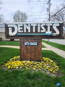 Weathered wood monument sign reading "Dentists" with a phone number panel, surrounded by yellow and white flowers, before replacement by Adams Signs and Graphics