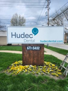 Newly installed Hudec Dental monument sign with logo, website URL, and phone number panel on a wood post base, photographed from the front during installation by Adams Signs and Graphics