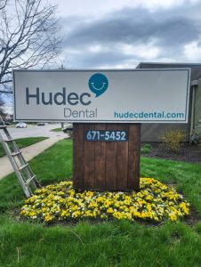 Side-angle view of the completed Hudec Dental monument sign installation showing the white panel with teal logo and hudecdental.com URL, mounted on a wood post base with a flower bed at the base
