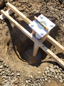 Overhead view of a sign post base plate with anchor bolts and a spirit level resting on top, set in an excavated hole with wood bracing holding the post plumb before concrete is poured | Adams Signs & Graphics