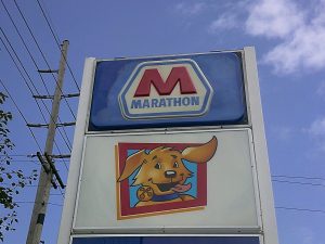 A close view of a Marathon pole sign totem showing the illuminated M logo panel above the Speedy dog mascot panel, framed by trees and a partly cloudy sky | Adams Signs & Graphics