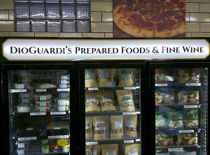 Backlit interior sign reading DioGuardi's Prepared Foods and Fine Wine mounted above deli freezer cases stocked with pasta and cannoli | Adams Signs & Graphics
