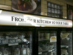 Backlit interior sign reading From Our Kitchen To Your Table above deli cases holding fresh meatballs and garlic bread | Adams Signs & Graphics