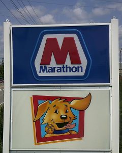 A tall pole sign totem with an illuminated Marathon logo panel above a Speedy dog mascot panel, set against a blue sky with power lines nearby | Adams Signs & Graphics