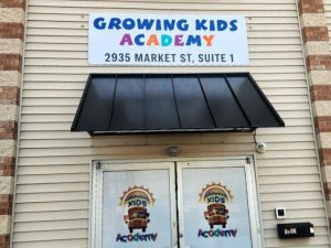 Building sign, black awning, and door graphics for Growing Kids Academy featuring the business name and a colorful school bus logo | Adams Signs & Graphics