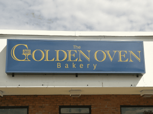 Blue backlit cabinet sign with gold lettering reading The Golden Oven Bakery and a crowned cake stand logo, mounted on a white storefront fascia | Adams Signs & Graphics