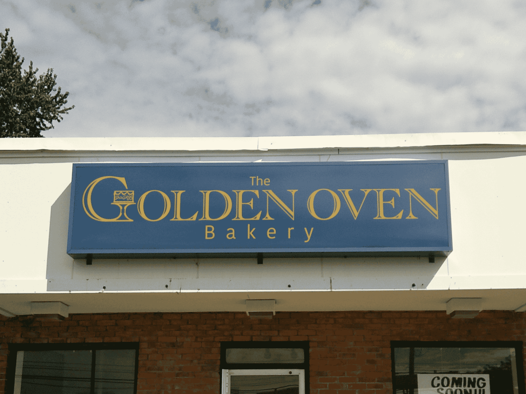 Angled view of the blue cabinet sign for The Golden Oven Bakery mounted on a white fascia beneath a cloudy sky | Adams Signs & Graphics