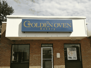 Brick bakery storefront with a blue and gold wall sign for The Golden Oven Bakery above the entrance and a coming soon poster in the window | Adams Signs & Graphics