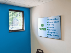 Angled view of a brushed aluminum sign listing the HealthMarkets promise values, mounted with polished standoffs beside a blue accent wall and window | Adams Signs